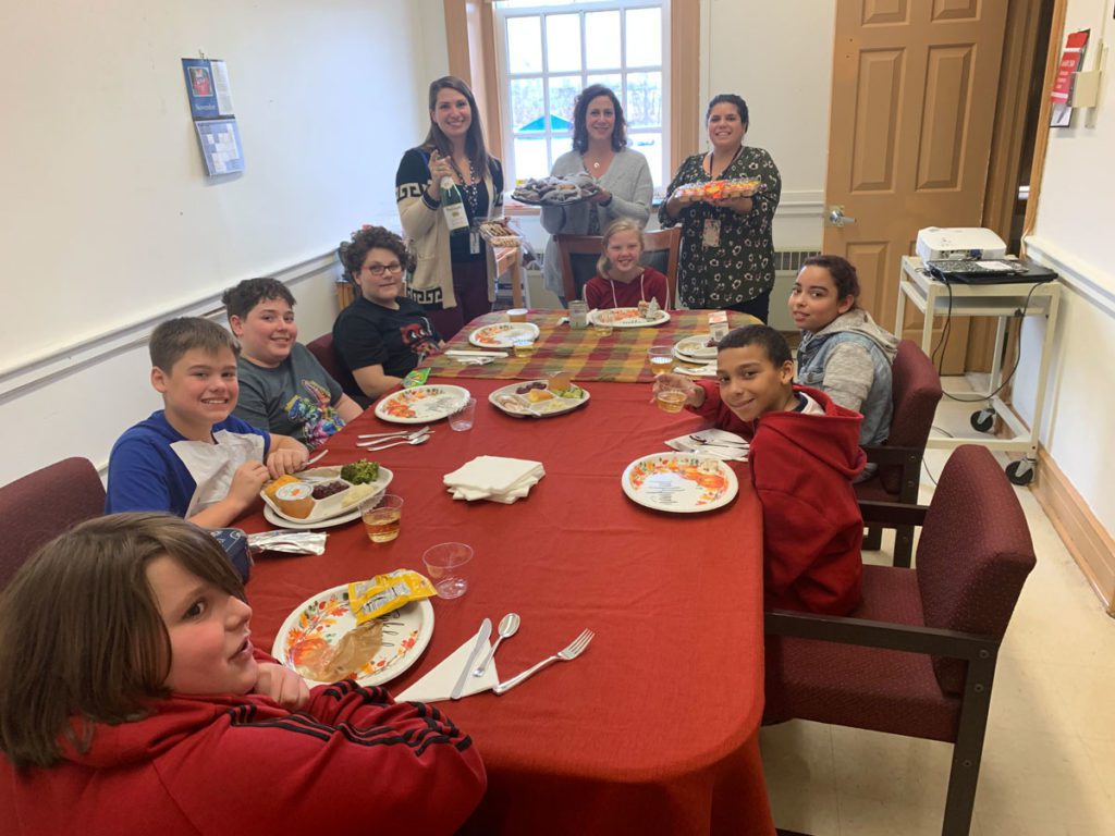 students eating turkey dinner at table while teachers hold up trays of desserts