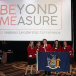 Four girls an bou standing on stage in FCCLA uniforms in from of banner that reads "Beyond Measure"