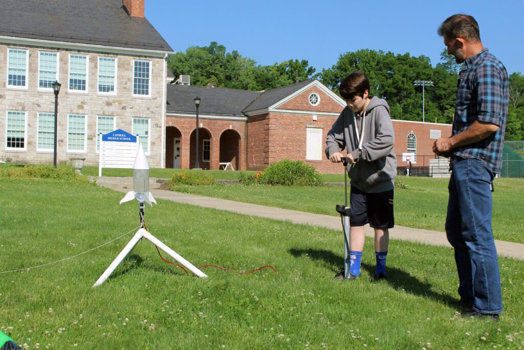 boy priming water rocket with bicycle pump outside CMS while man looks on