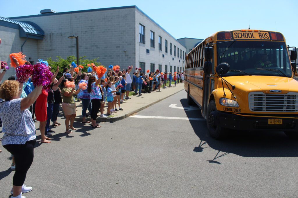 staff wave to buses leaving elementary school parking lot.