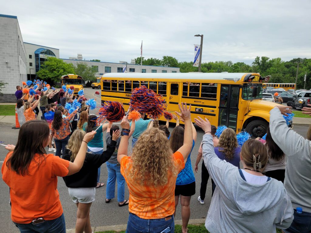 crowd of school staff waving and shaking pom-poms at busses
