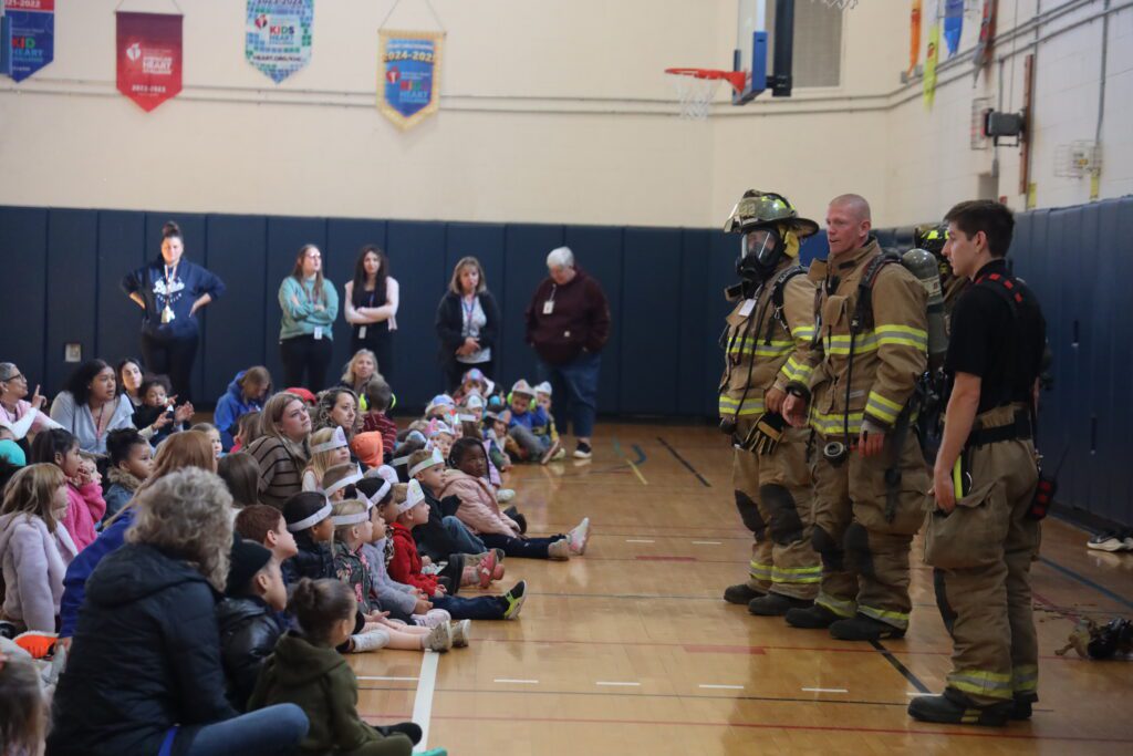 firiefighters in firefighting gear lead presentation to large audience of children