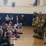 firiefighters in firefighting gear lead presentation to large audience of children