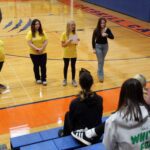 girls in yellow shirts talking to students seated on gym bleachers