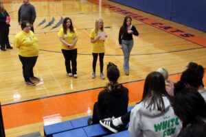 girls in yellow shirts talking to students seated on gym bleachers