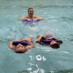 students swimming on their backs holding kickboards while being instructed by woman