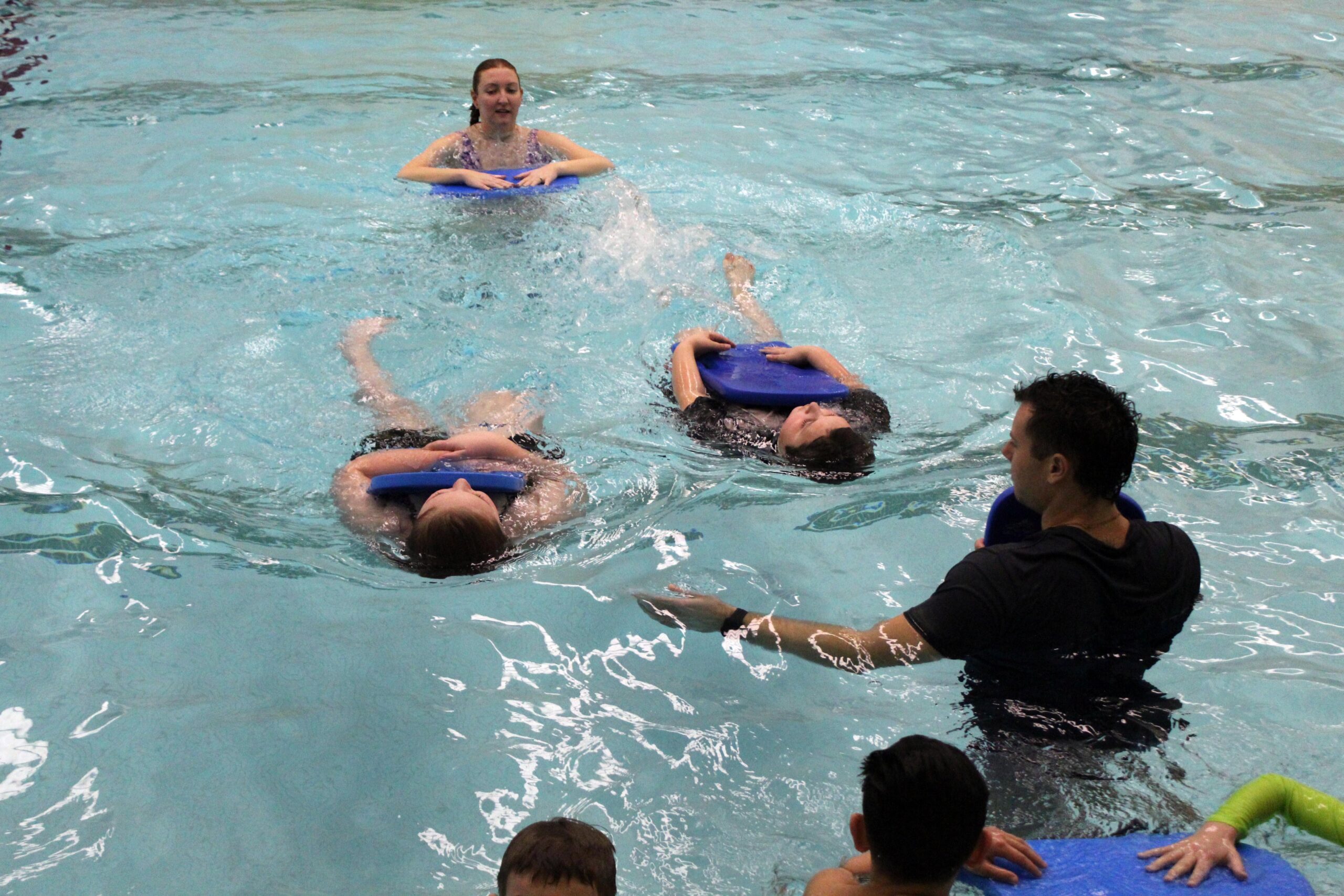 students swimming on their backs holding kickboards while being instructed by man and woman