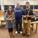 man flanked by woman holding saxophone and man holding trombone.