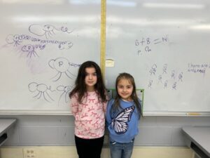 two girls posing next to math problem on white board