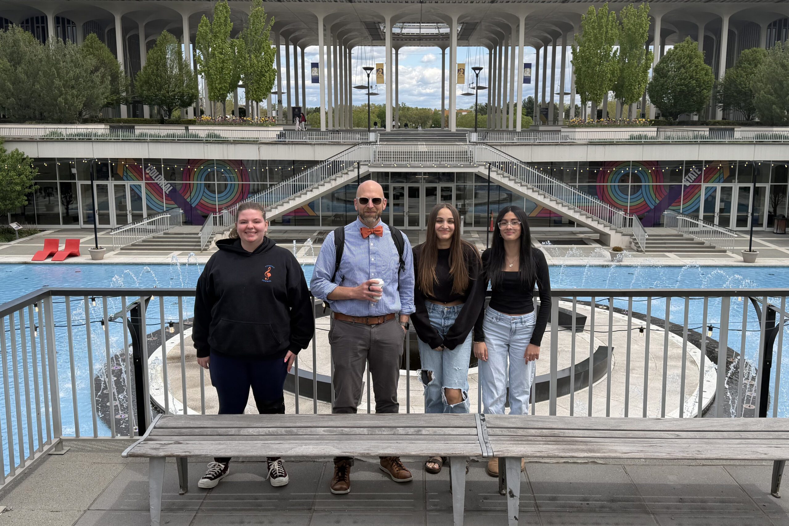 three young women and man in bowtie pose for photo on UAlbany campus